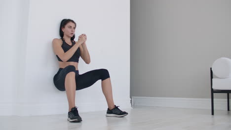 The-brunette-girl-froze-in-a-squat-leaning-on-the-wall-doing-a-static-exercise-in-a-bright-interior.-Keep-90-degree-leg-angle